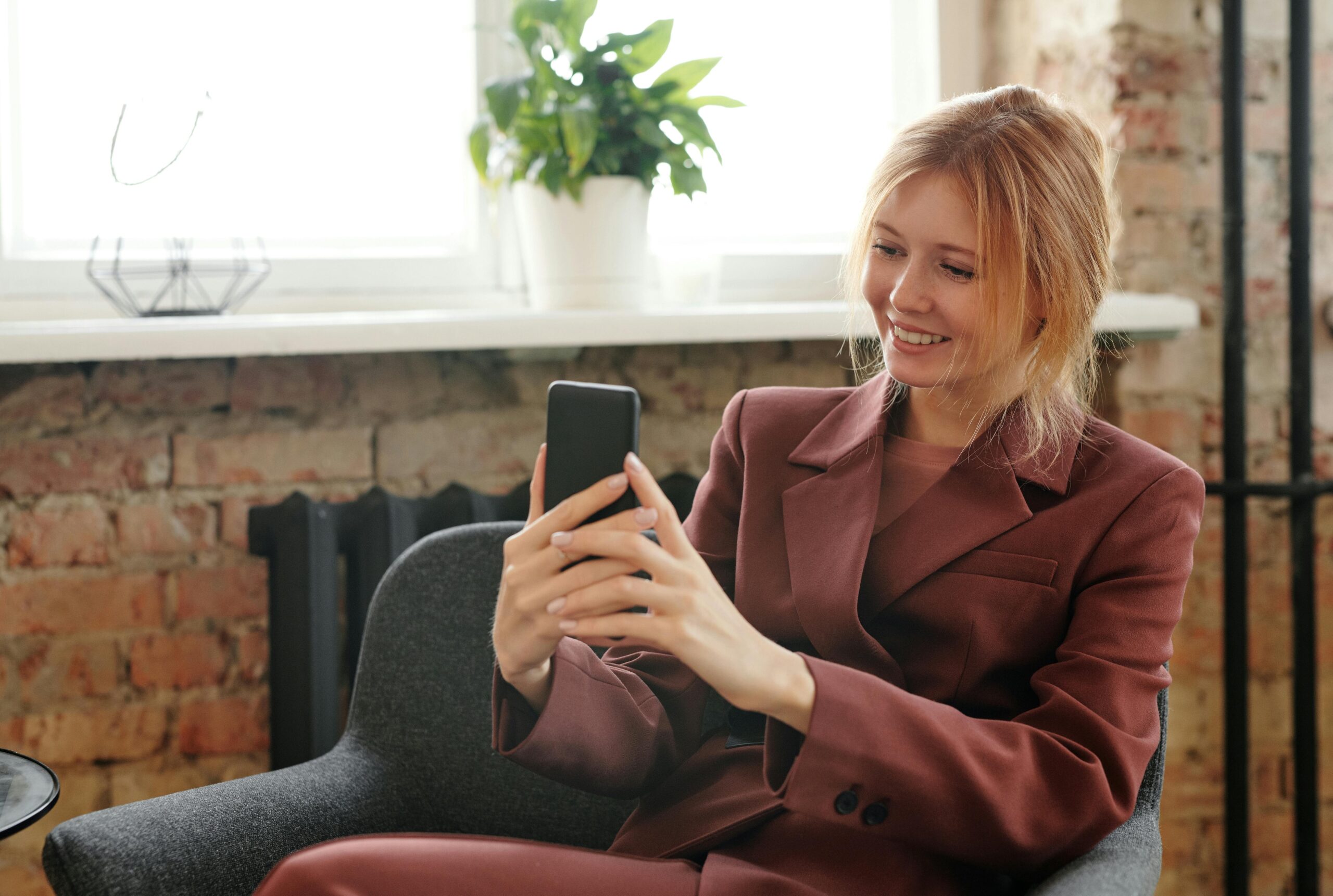 Woman in a stylish blazer sitting on a chair using a smartphone indoors, capturing a happy moment.